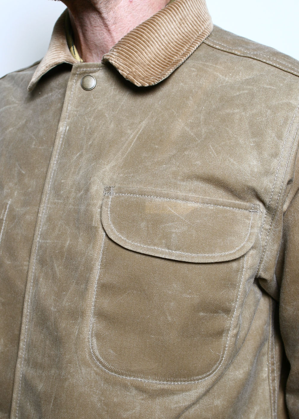 Close detail of a Rogue Territory Supply Jacket in tan waxed canvas, showing the corduroy collar, snap placket, and a rounded chest patch pocket, the waxed cloth marked with natural creasing